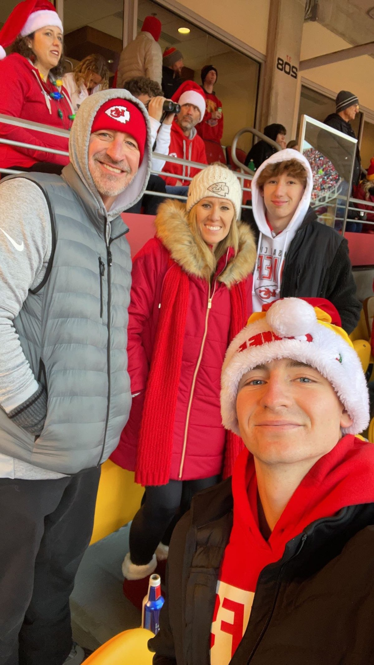 Four people in Kansas City Chiefs red attire and Santa hats pose together indoors at what appears to be a stadium or sports venue.