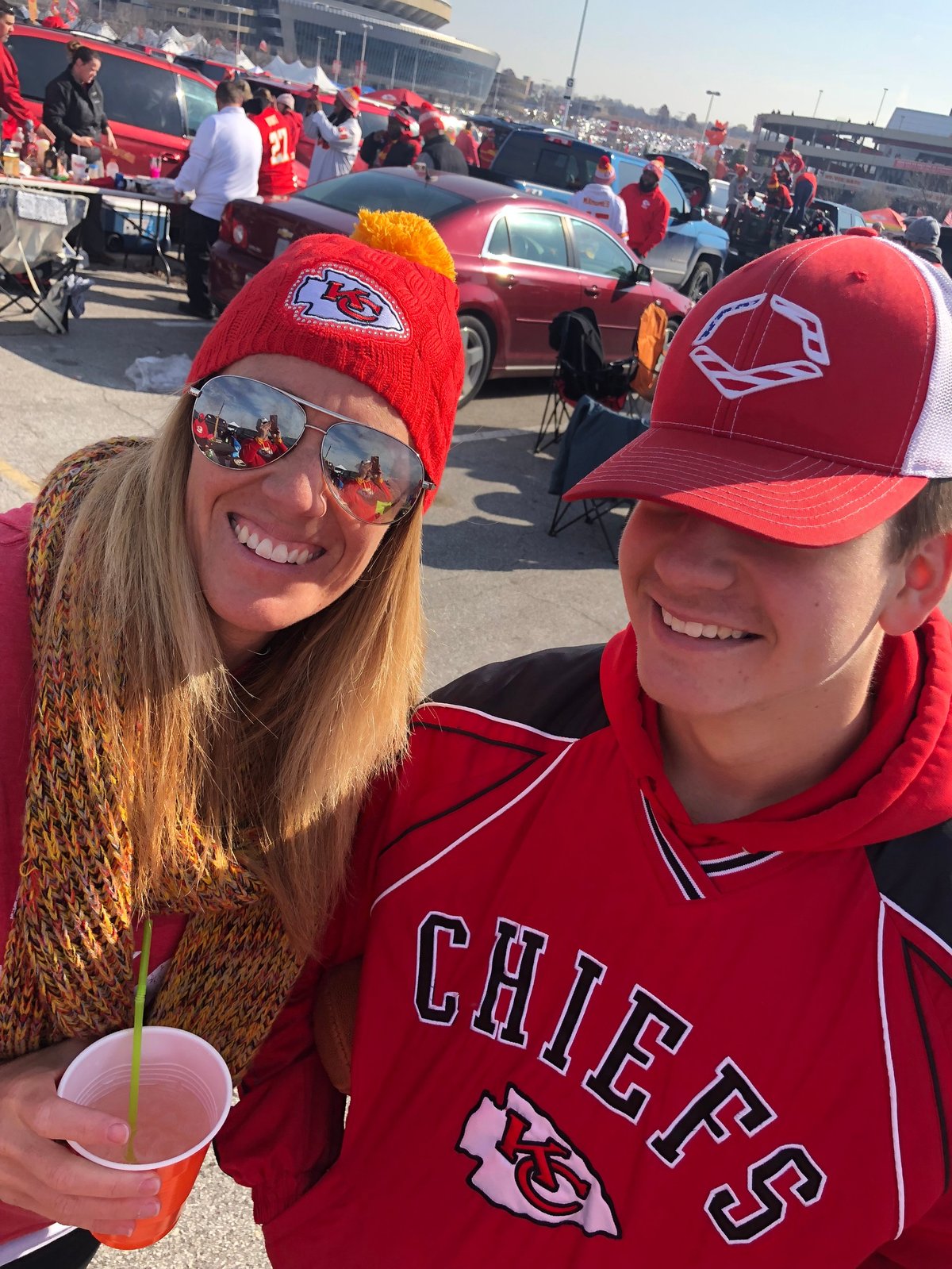 Two fans wearing red Kansas City Chiefs gear smile at a tailgate party with cars and crowd in background