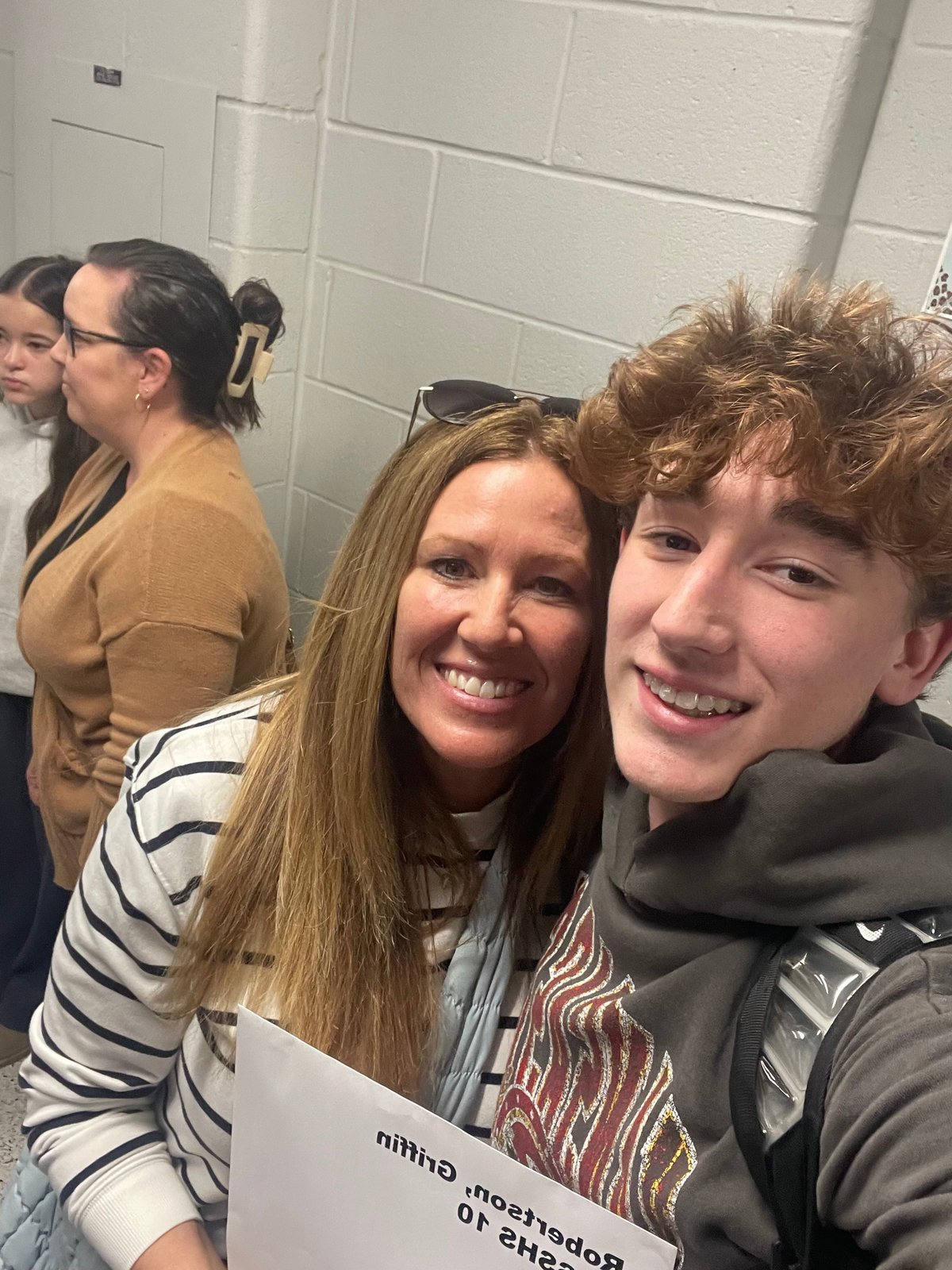 A woman and teenage boy smile at the camera in a school hallway, with other students visible in the background near white subway-tiled walls.
