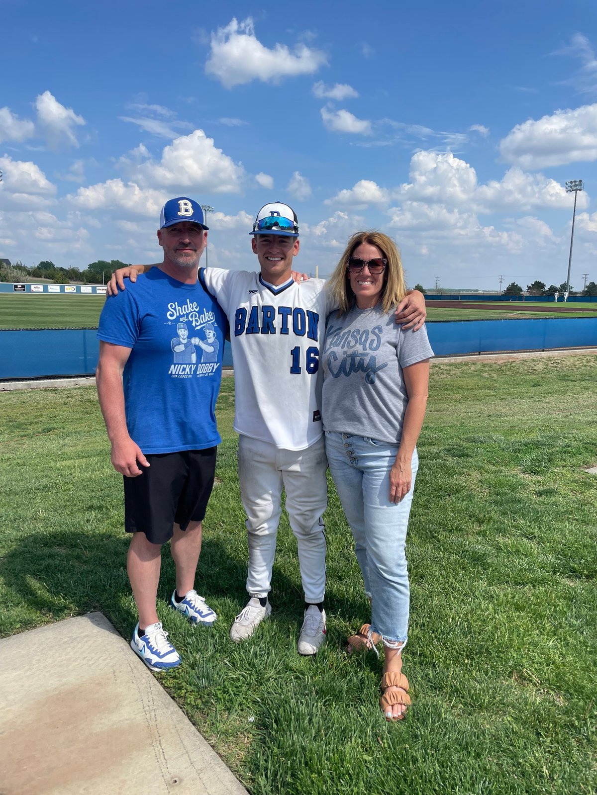 Three people posing together at a baseball field with blue sky and white clouds in background