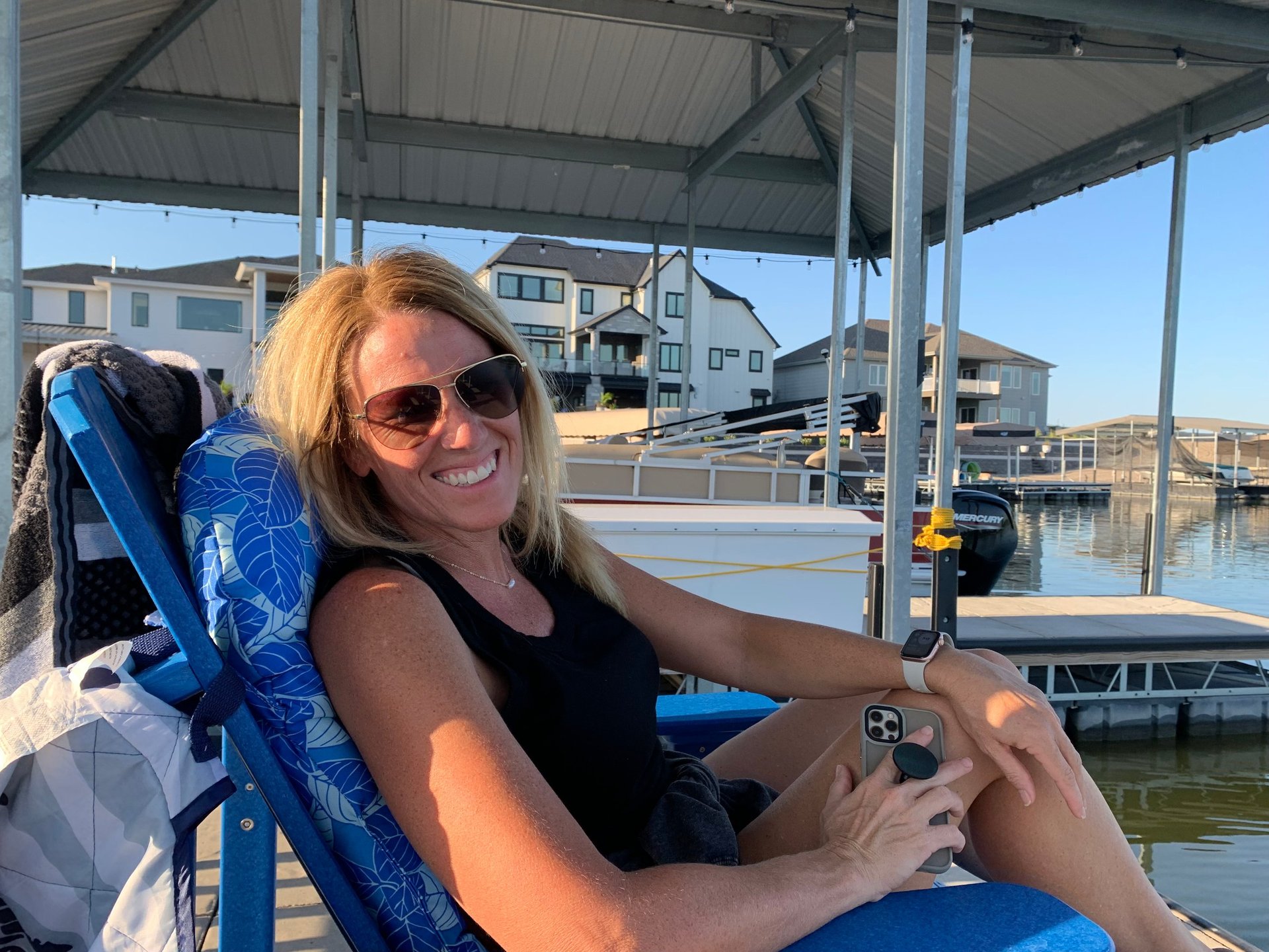 Woman in sunglasses smiling at the helm of a boat docked near waterfront houses on a sunny day