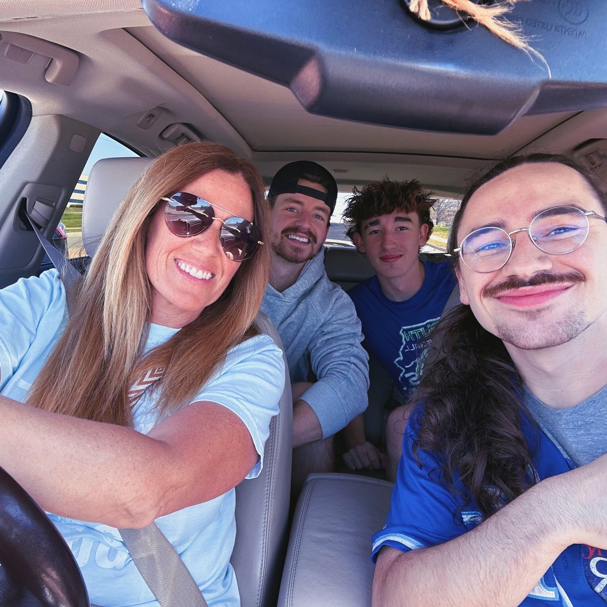 Four friends smiling and taking a selfie inside a car, all wearing casual clothing and sunglasses