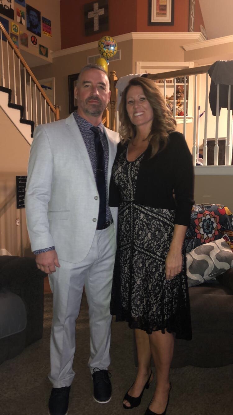 Man in light blue suit and woman in black patterned dress standing together indoors with holiday decorations and staircase in background