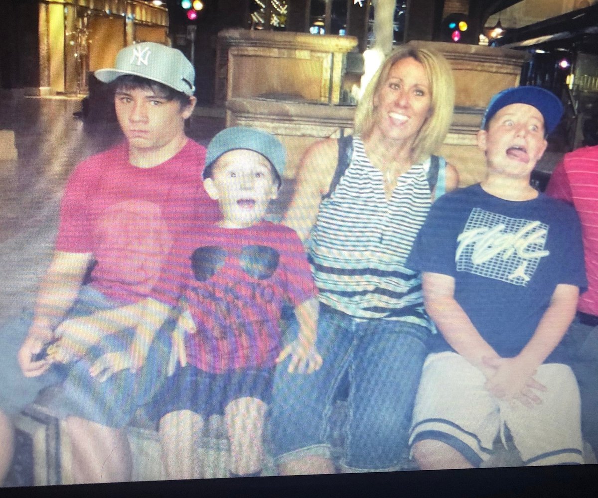 Woman with three young boys posing together indoors. They are wearing casual clothing and baseball caps. Warm indoor lighting in background.
