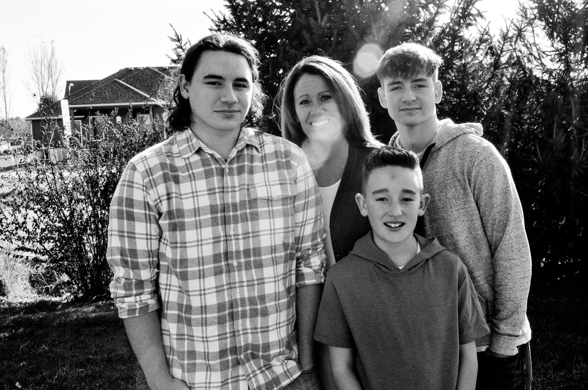 Black and white photo of four smiling people posing outdoors with trees and a house in the background