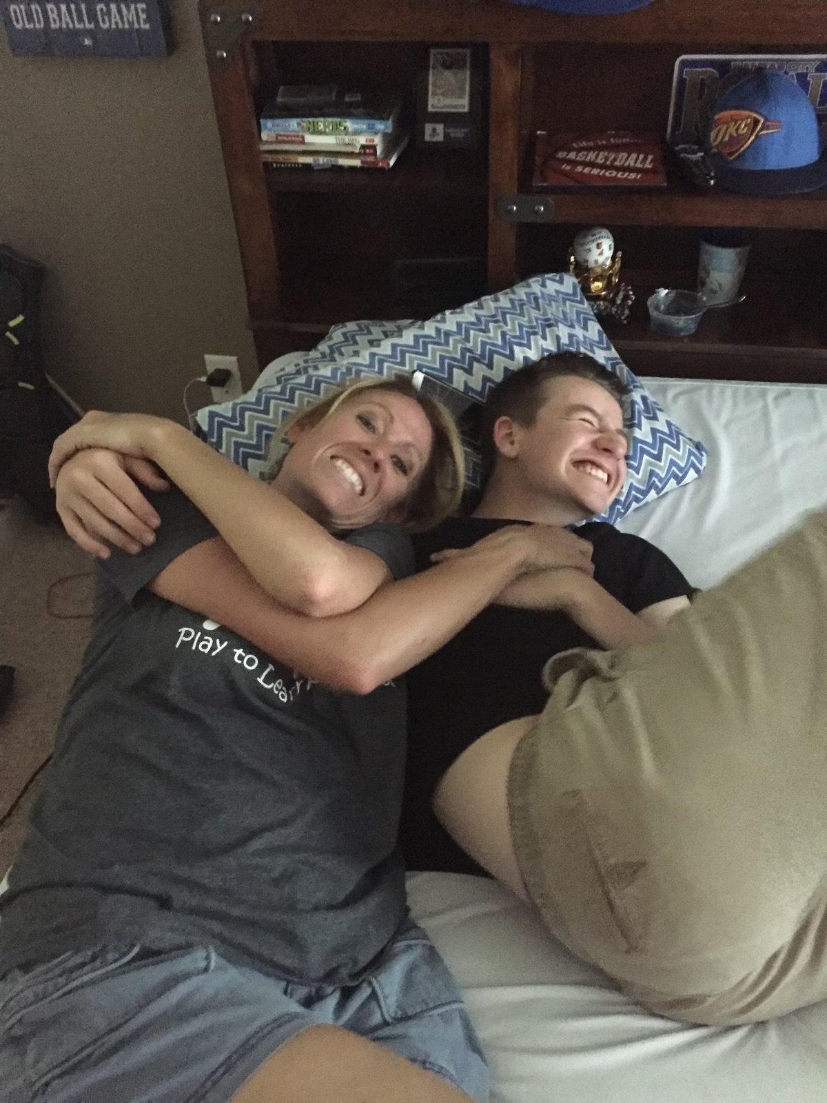 Two people smiling and embracing on a bed in a bedroom with sports memorabilia and wooden furniture visible in the background