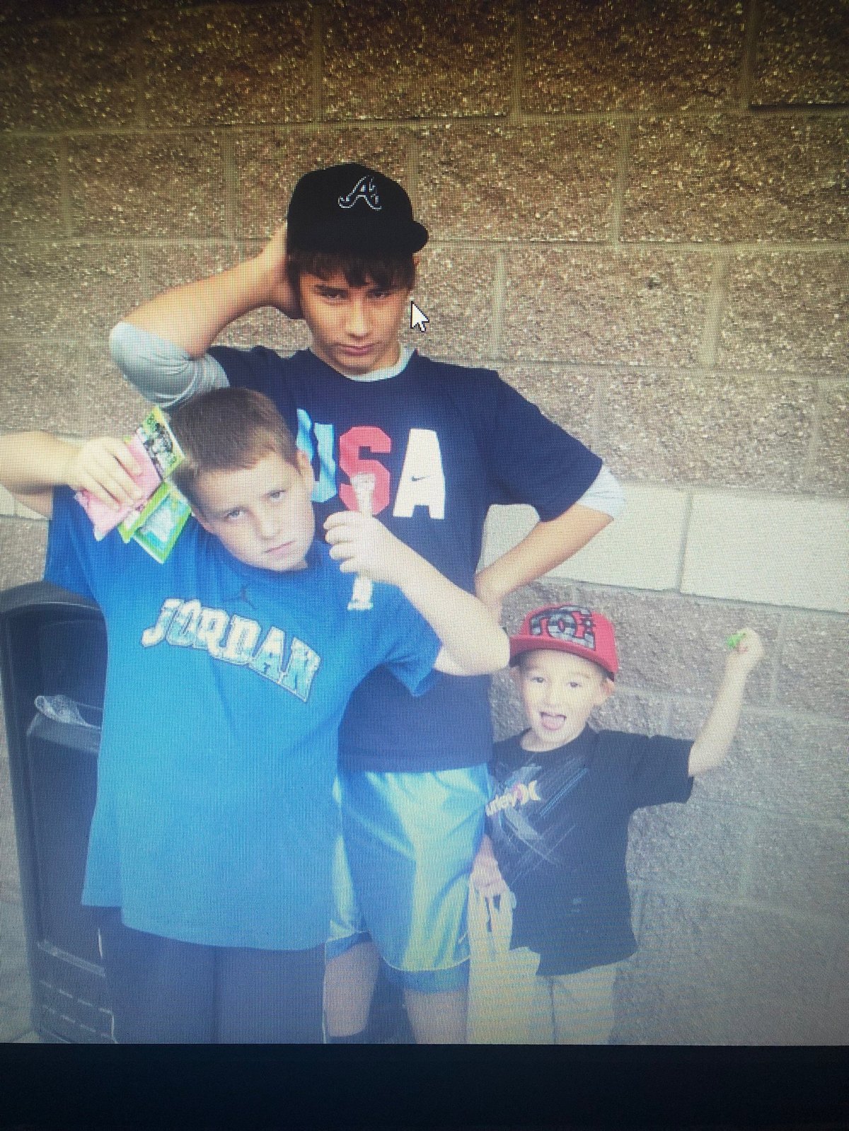 Three young boys in athletic shirts posing together against a brick wall, wearing baseball caps and casual sports apparel