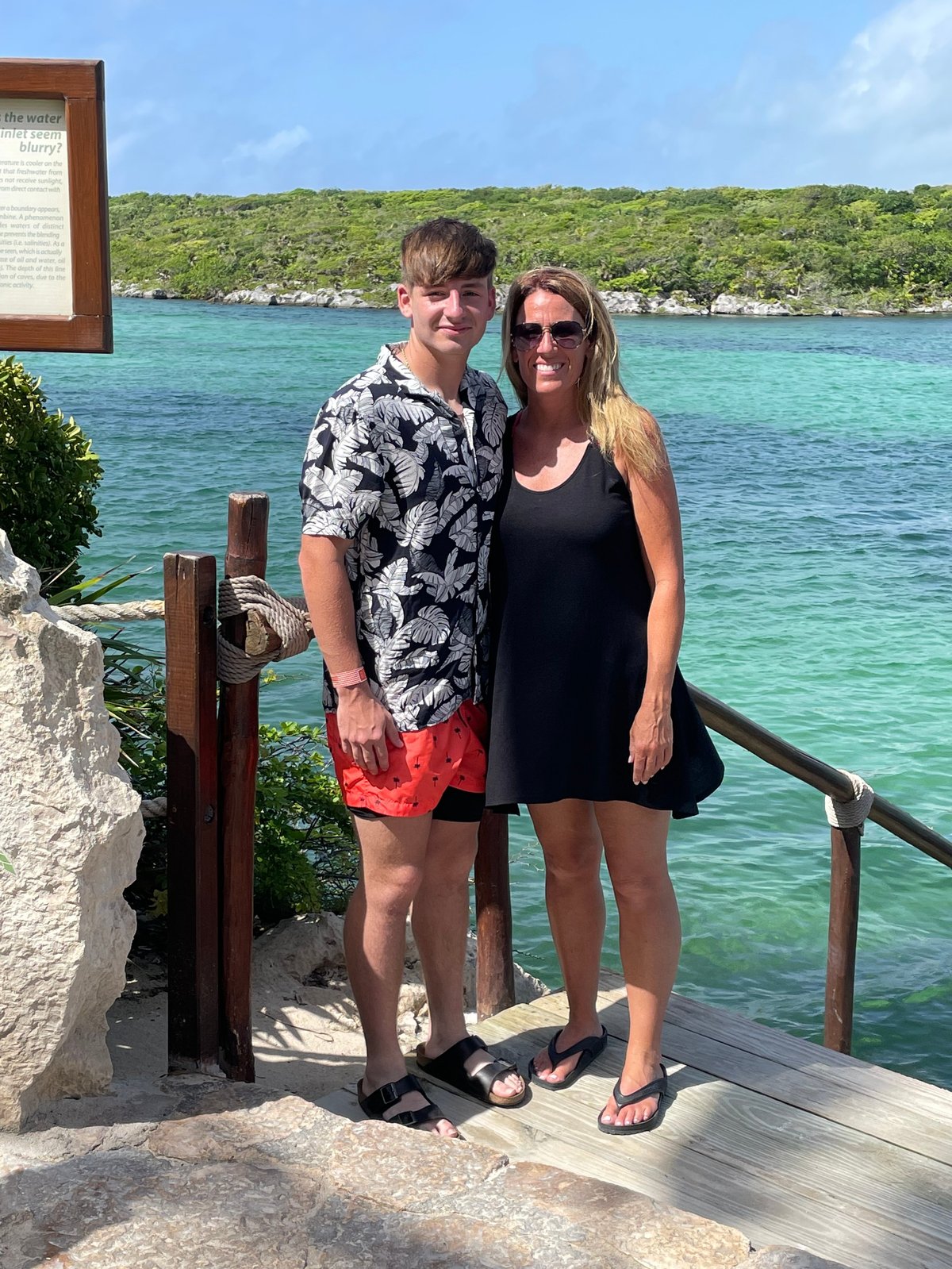 Two people posing together on a wooden deck by a turquoise tropical beach with lush green hills in the background