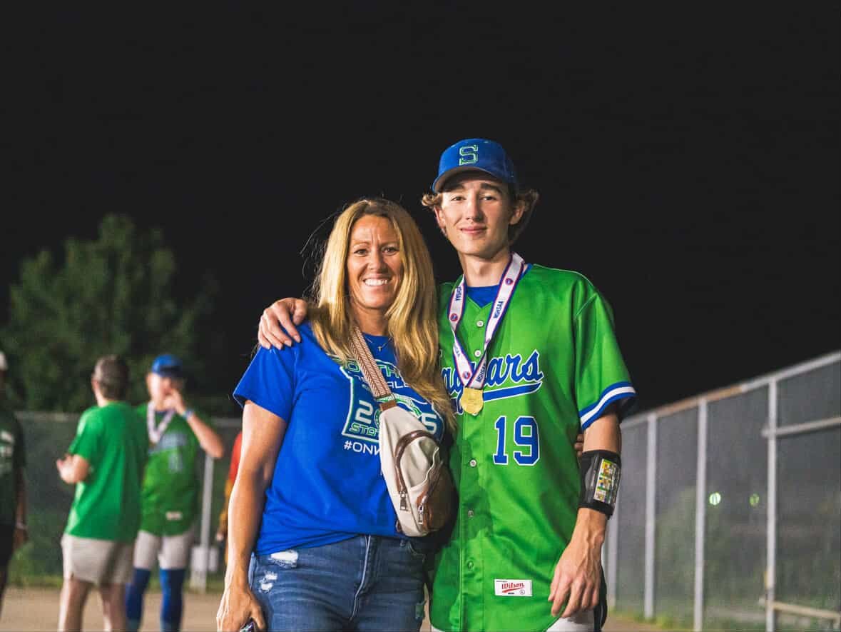 Young man in green baseball uniform wearing medals standing with woman in blue shirt on athletic field