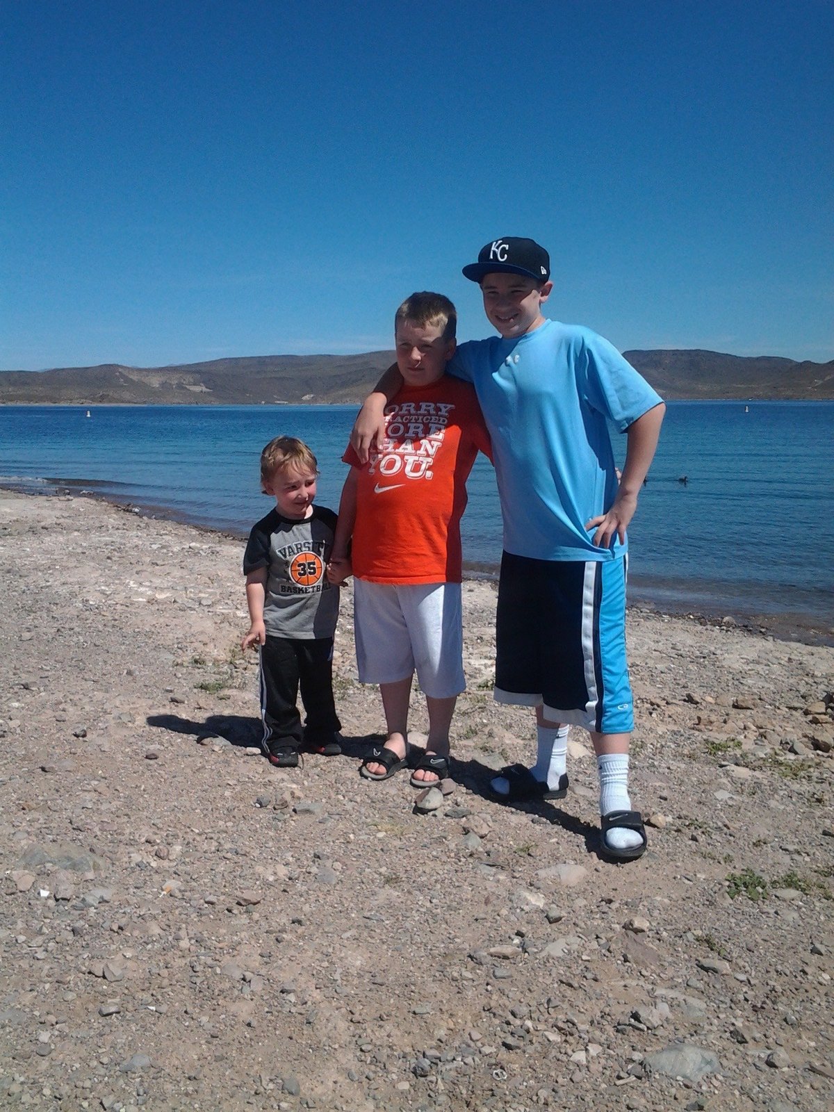 Three children standing on a sandy beach with calm turquoise water and mountains in the background under clear blue sky